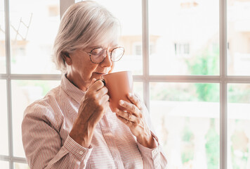 Portrait of sad pensive senior woman at home close to the window holding a cup of tea lost in her thoughts