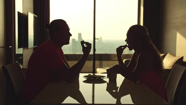 Silhouette cute family couple sitting at kitchen table in home. Husband and wife in morning drinking coffee. Window city view. Man and woman together spend weekend, eating cakes. Food and beverages