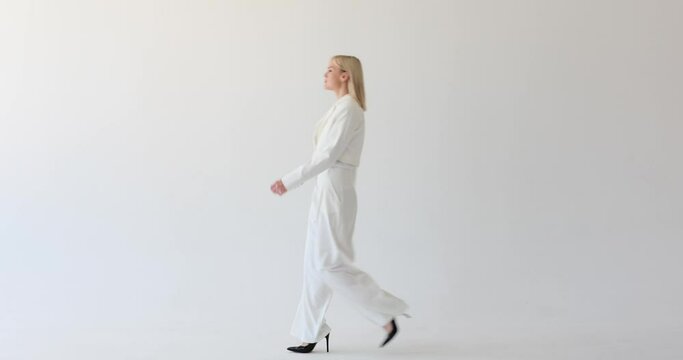 A blonde woman is seen on a clean white backdrop, dressed in a professional suit, walking by with a purposeful stride. Her confident and composed demeanor indicates her sense of purpose as she moves.