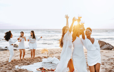 Carefree ladies bride team toasting wine glasses while having hen party and dancing on the beach at...