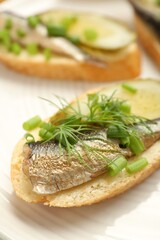 Delicious sandwich with sprat, pickled cucumber, green onion and dill on plate, closeup