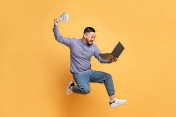 Big Profit. Excited Young Asian Man Jumping With Laptop And Dollar Cash
