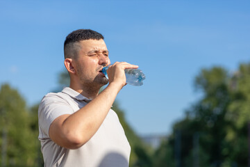 adult man drinking water from bottle in heat. guy suffering from thirst in against blue sky. prevention of dehydration on sunny day. thirsty male drink liquid for hydration, wellness, health