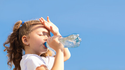 little girl drinking water from bottle in heat. kid suffering from thirst in against blue sky....