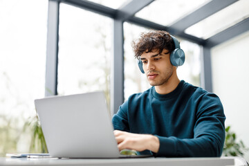 Young european male designer listening music on headphones while working on laptop at a desk in...