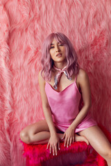 Happy young sexy Asian woman with pink wig hair and a short satin dress sitting on a chair a fur background in the studio. Fashion