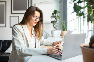 Positive businesswoman working with papers and laptop in office, female entrepreneur sitting at desk, free space