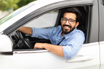 Cheerful eastern guy with glasses driving brand new white auto