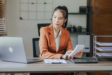 Portrait of Asian businesswoman in suit calculating company sales with calculator Laptop, smart phone and tablet on table