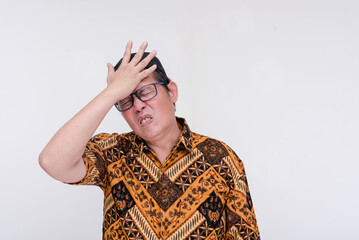 An indonesian man slaps his forehead, realizing he forgot something. Annoyed by his absentmindedness or failure. Wearing a batik shirt and songkok skull cap. Isolated on a white background.