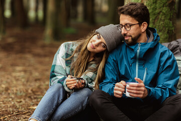 Fototapeta premium Relaxed happy young caucasian couple in jackets with cups of tea from thermos, enjoy travel, vacation, rest