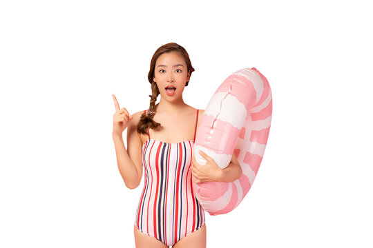Happy young Asian woman wear swimsuit hold pink swim ring or inflatable tube ring Excited lady pointing finger up to copy space isolated on pink color background studio Summer vacation concept