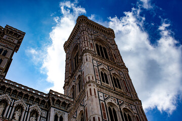 Looking skyward at Giotto's Bell Tower, adjacent to Florence Cathedral, its verticality is emphasized by the cloud-streaked blue sky, showcasing its ornate Gothic architecture. - obrazy, fototapety, plakaty