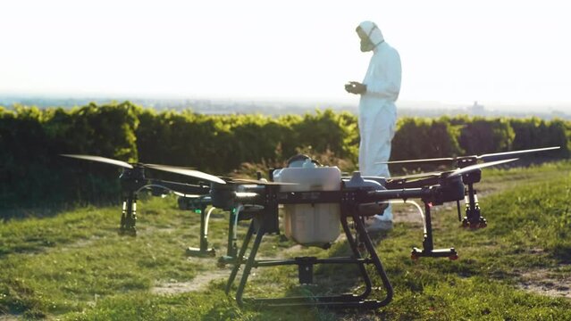 Slow motion footage of an agricultural drone taking off to spray plants with an engineer in a protective suit in the background controlling the aircraft