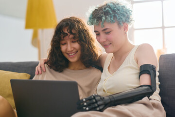 Happy young woman with disability and her girlfriend watching online video while sitting on couch at home and looking at screen of laptop