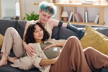 Happy young pregnant woman and her girlfriend with myoelectric hand relaxing on comfortable couch...