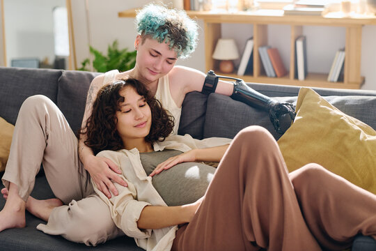 Happy young pregnant woman and her girlfriend with myoelectric hand relaxing on comfortable couch with cushions in home environment