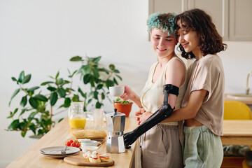 Young affectionate woman embracing her girlfriend with myoelectric arm holding cup of coffee while...