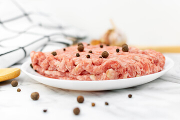 Fresh raw minced meat with peppercorns on a white plate and ingredients for cooking on a marble table. Uncooked ground beef, pork.
