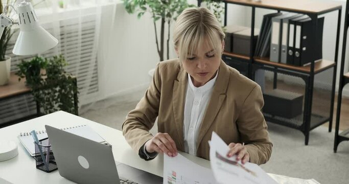 Serious and focused businesswoman is depicted meticulously comparing information in documents with her laptop. With a determined expression, she meticulously cross-references data.