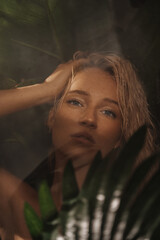 Close-up portrait of an enchanting blonde in a black swimsuit behind a shower glass surrounded by palm leaves, tropical concept, vacation concept in tropical countries