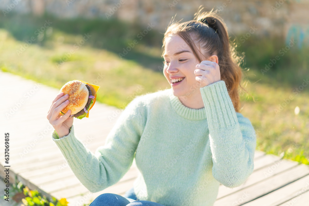 Wall mural Young pretty girl holding a burger at outdoors celebrating a victory