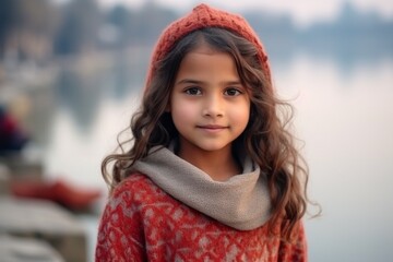 Portrait of a beautiful little girl with long curly hair in a red knitted hat and scarf on the background of the lake