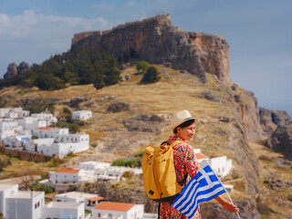 Fototapeta na wymiar Young traveling woman with national greek flag enjoying view of Lindos. Travel to Greece, Mediterranean islands outside tourist season.