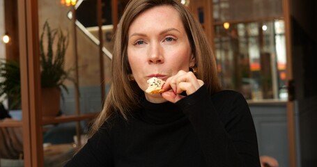 Beautiful stylish woman sit in restaurant bite piece of seeded farm white bread. Medium shot of female adult in black shirt with long sleeves eat fresh crispy bun with sesame.