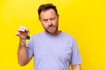 Middle age man holding home keys isolated on yellow background with sad expression