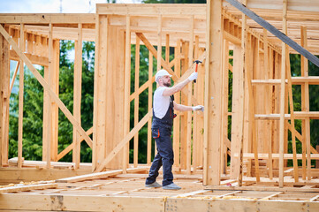 Carpenter constructing wooden frame, two-story house near forest. Bearded man hammering nails with...