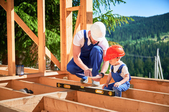 Father with toddler son building wooden frame house. Male worker showing his son the construction plan, wearing helmets and blue overalls on sunny day. Carpentry and family concept.
