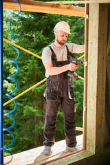 Carpenter constructing wooden framed house. Bearded man worker cladding facade of house, fastening with screwdriver, wearing work overalls and helmet. Concept of modern eco-friendly construction.