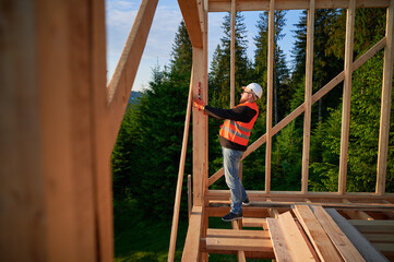 Carpenter constructing wooden frame house. Bearded man, wearing spectacles, inspects the walls for...