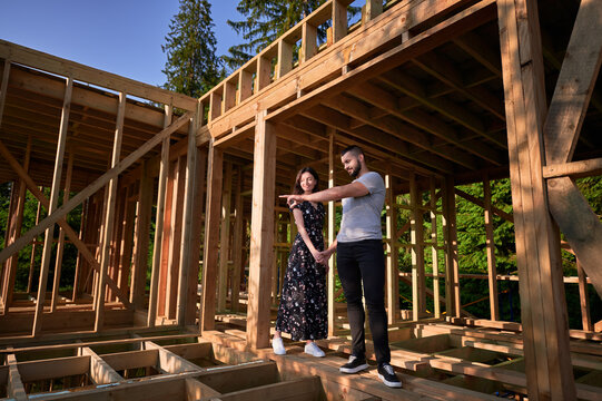 Man and woman inspecting their future wooden frame dwelling nestled near forest. Youthful couple at construction site in early morning. Concept of contemporary ecological construction.