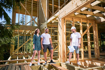 Man with detailed plan shows to couple the process of constructing wooden framed house. Happy...