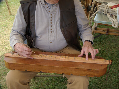 The builder of this dulcimer plays a tune, using a feather to pluck the strings while he chooses the notes on the fret with his other hand.