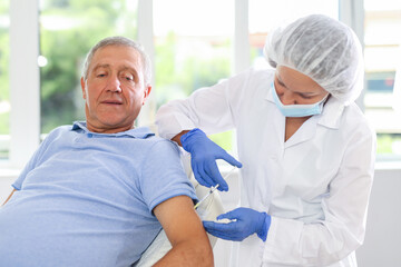 Female nurse in disposable cap and mask gives injection, injects vaccine into shoulder of elderly male patient of clinic