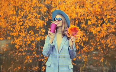 Autumn portrait of beautiful happy smiling woman with yellow maple leaves and cup of coffee wearing round hat and coat in the park