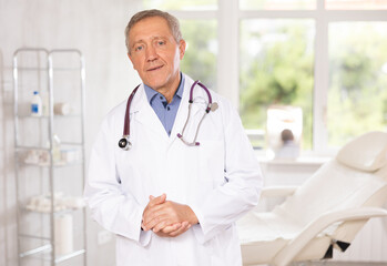 Positive old man doctor posing against background of doctor's cabinet with clinical chair