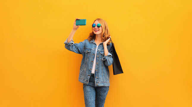 Beautiful happy smiling woman taking selfie with mobile phone with shopping bags wearing denim jacket on yellow studio background