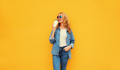 Beautiful caucasian smiling young blonde woman with cup of coffee wearing denim jacket on yellow background
