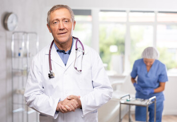 Fototapeta premium Experienced smiling elderly male doctor in white coat standing in modern light medical office with female assistant in blue uniform preparing instruments in background