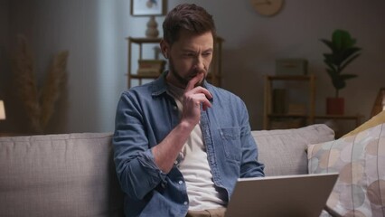 Man working late in evening. Male sitting at couch and using tablet. Thoughtfull man working distantly. Bearded man wearing wireless headphones. Serious man resolving business issues.