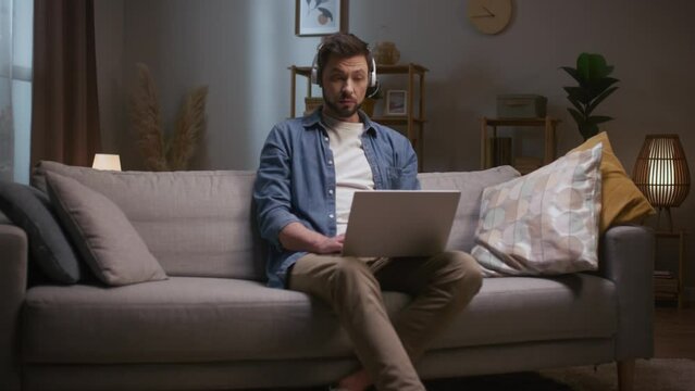 Man looking at screen and talking. Male wearing headphones. Man conducting video conference. Man using laptop and headset. Male sitting at couch at living room. Male joining online meeting.
