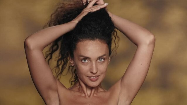 A brunette woman ties, twists her thick long curly hair into a bun. Portrait of a smiling seminude woman in the studio on a yellow background with highlights. The concept of beauty, hair styling, care