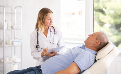Middle-aged woman physician making notes while discussing treatment procedures with male patient lying nearby
