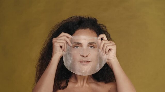 A woman applies a cosmetic white sheet mask on her face, smoothes it. Portrait of a seminude woman in the studio on a yellow background. The concept of beauty, cosmetology, care.