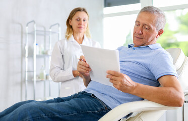 Fototapeta premium Before medical manipulation, mature man patient sits in chair and reads documents with woman doctor about upcoming manipulation. Medical documentation, questionnaire, patient survey