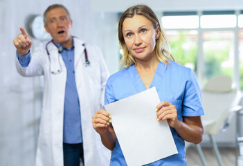 Middle-aged nurse standing with sad look in cabinet while angry mature doctor is scolding her behind
